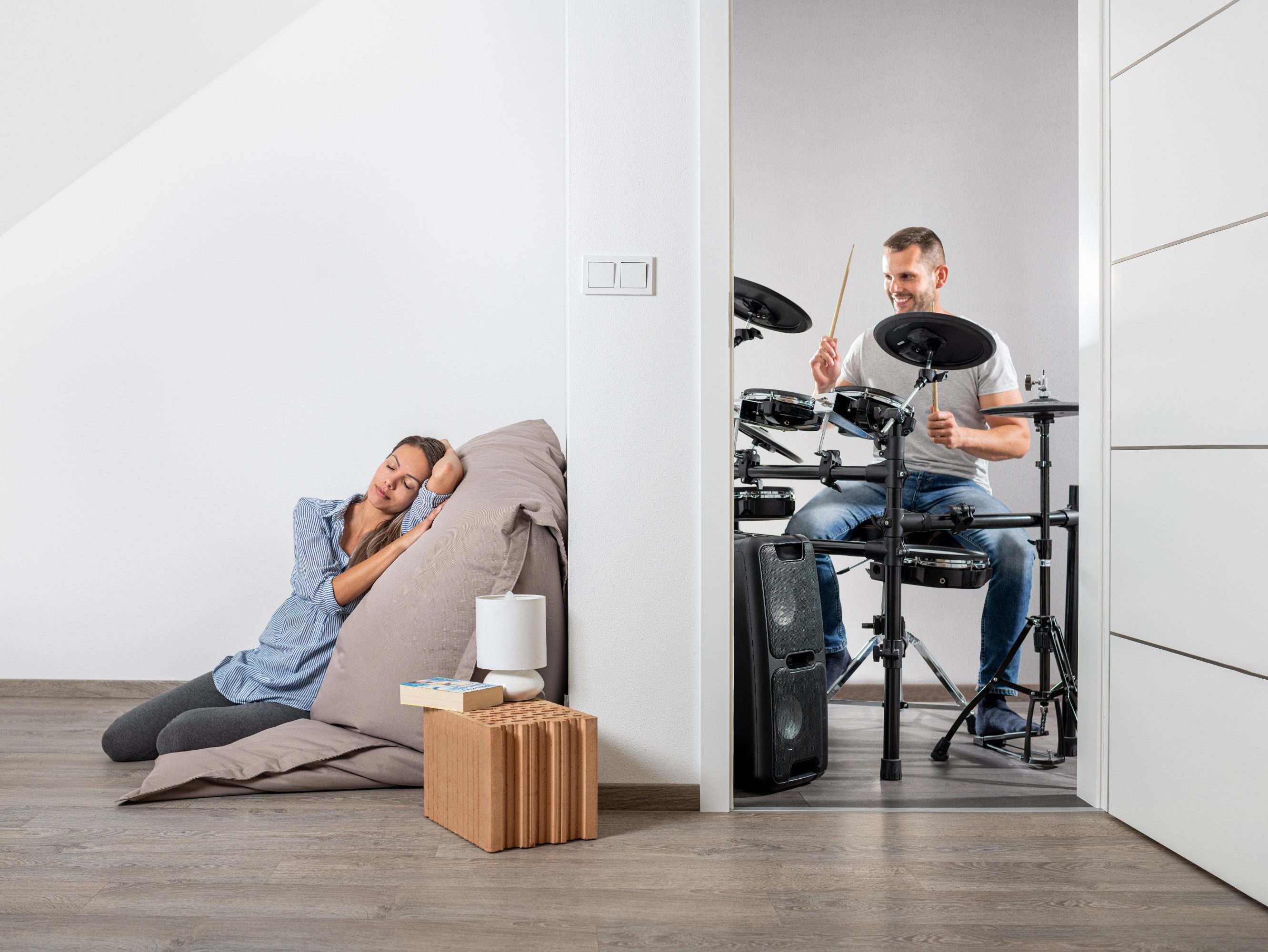 Muž hrající na bicí a v druhé místnosti klidně ležící žena cz